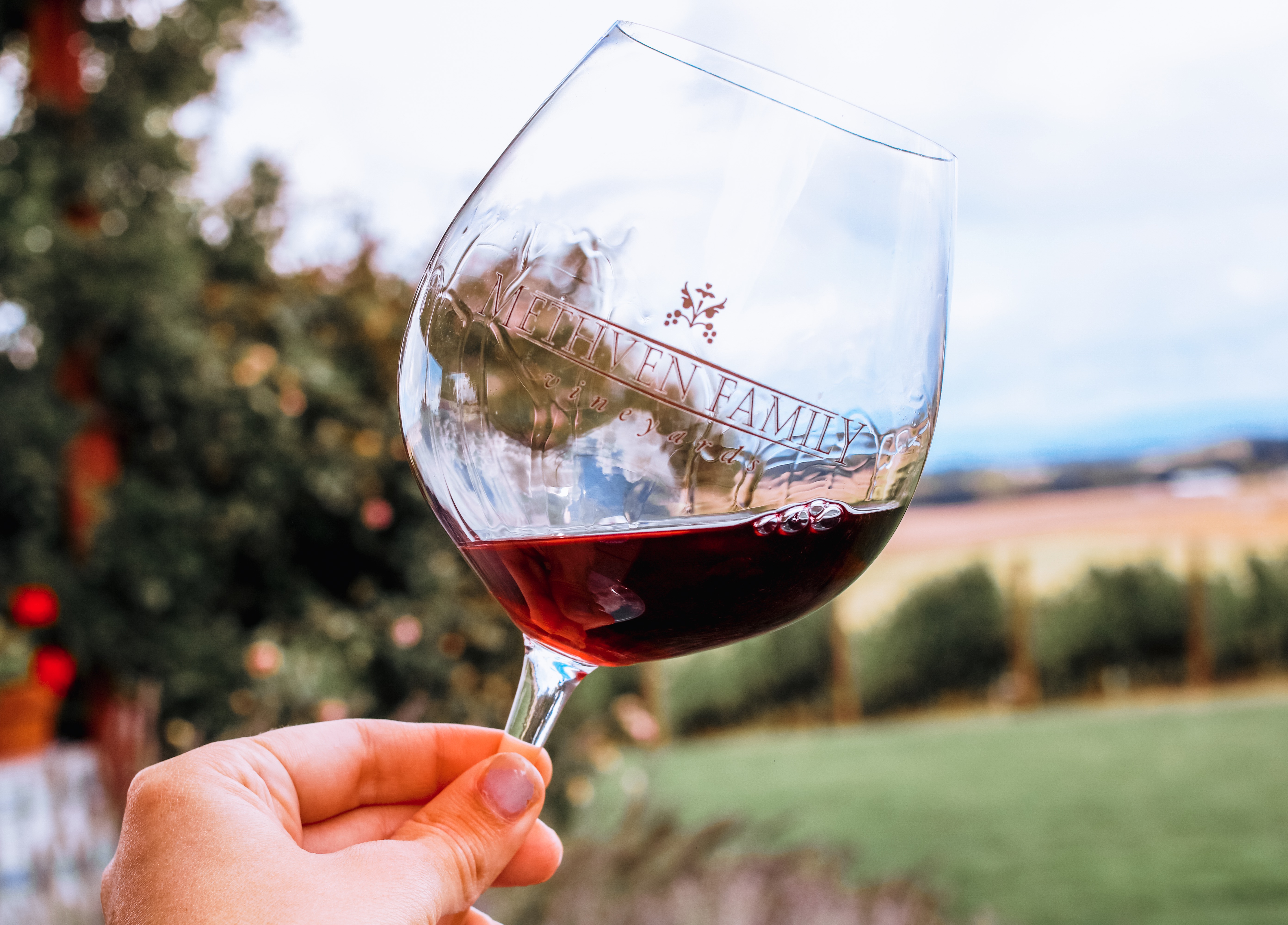 In the foreground, a Methven Family Vineyard branded wine glass, filled part-way with a red pinot, tilted at an angle. In the background, an unfocused view of our estate vines and some evergreens on the edges of the picture.
